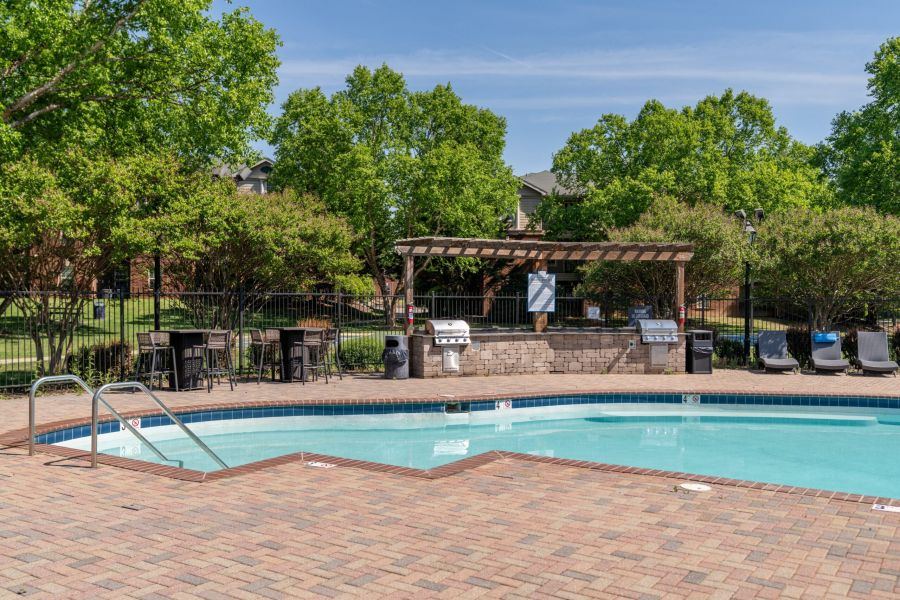 Attain on 5th apartment homes with Outdoor swimming pool with brick deck, lounge chairs, and barbecue grills under a wooden pergola, surrounded by trees.
